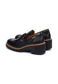 Mocasines de cuero negro con suela baja tipo plataforma, que presentan una costura marrón contrastante y un acento decorativo en la parte superior.