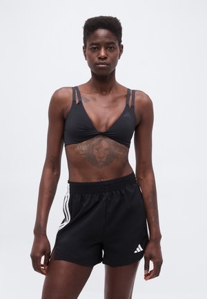 Atletisk person iført sort sports-bh og sorte Adidas-shorts med hvide striber, stående mod en ensfarvet lys baggrund.