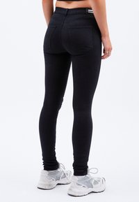 Schwarze Skinny Jeans mit hohem Bund, ausgestattet mit zwei Gesäßtaschen und satinierter Oberfläche. Kombiniert mit weißen Sneakers.