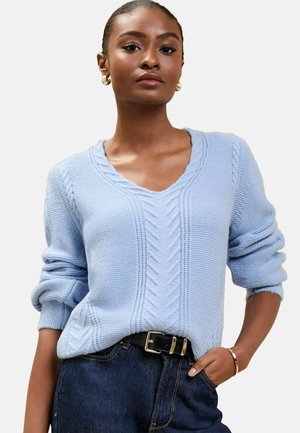 Hellblauer Strickpullover mit V-Ausschnitt, der Kabelstrickmuster und umgeschlagene Ärmeln aufweist. Kombiniert mit dunkler Jeans und einem schwarzen Gürtel.