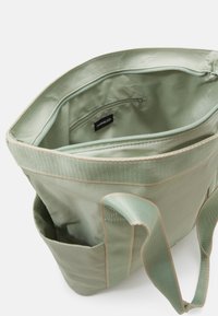 Sac fourre-tout en tissu vert avec une poche intérieure zippée. Présente une doublure vert clair et des coutures beiges contrastantes, ainsi que des poignées solides.