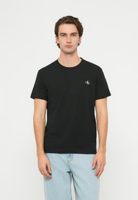 Čierne bavlnené tričko s okrúhlym výstrihom a krátkymi rukávmi, s malým bielym logom na ľavej strane hrudníka. Textúrovaný povrch.