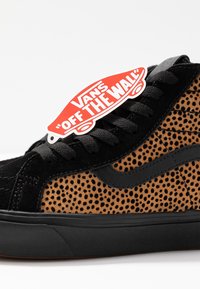 Baskets Vans en daim noir et marron avec des pois noirs, des lacets noirs et une étiquette rouge Vans « Off The Wall » accrochée près des lacets.