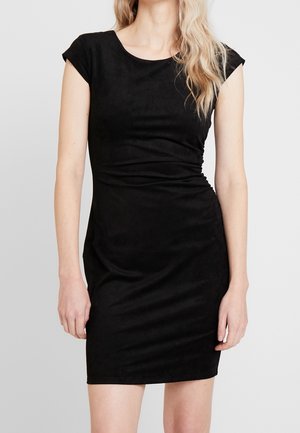 Frau mit langen blonden Haaren, die ein eng anliegendes schwarzes Kleid mit kurzen Ärmeln trägt, dargestellt vom Hals bis zur Mitte der Oberschenkel vor einem schlichten hellen Hintergrund.