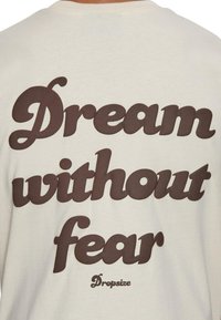 Lys beige t-shirt med stort mørkebrunt tekst, der står "Drøm uden frygt" og et mindre "Dropsize" logo nederst.