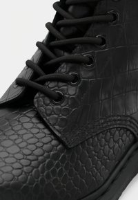 Gros plan d'une chaussure en cuir noir avec une texture en relief imitation peau de reptile et des lacets noirs passés dans des œillets métalliques.