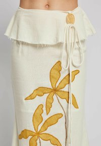 White linen skirt with a ruffled hem, featuring yellow embroidered palm leaves and a drawstring closure with a circular button detail.
