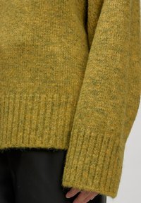 Mosterdgroene gebreide trui met geribde manchetten en zoom. Zachte textuur, licht oversized ontwerp, met een subtiel patroon overal.