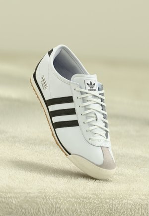 Baskets blanches avec des rayures noires, en cuir, dotées d'une semelle en caoutchouc texturée, d'un bout arrondi et d'un design à lacets. Marque Adidas visible.