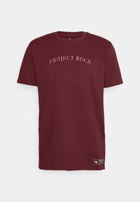 Camiseta cuello redondo de color burdeos con "PROJECT ROCK" bordado en un diseño curvado, con un pequeño parche de logo en el dobladillo. Material de algodón.