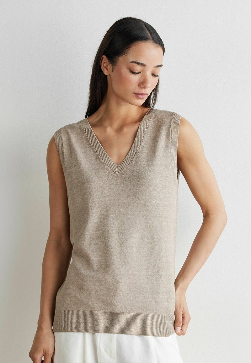 Femme portant un gilet tricoté beige sans manches à col en V, regardant vers le bas, debout contre un mur blanc uni.