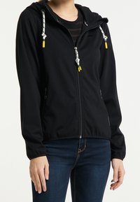 Personne portant un sweat à capuche noir zippé avec des cordons blancs et jaunes, et un jean bleu, debout devant un fond uni.