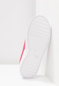 Semelle d'une basket Puma blanche et rose pressée contre un mur d'angle blanc et le sol, montrant le motif de la bande de roulement et le logo.