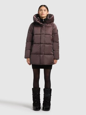 Pufferjakke i mørk mauve med hætte, quiltet design og lynlås foran. Båret sammen med en sweaterkjole og sorte knæhøje støvler.