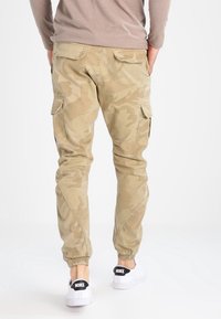 Jasnobeżowe spodnie bojówki w kamuflażu z zwężanymi mankietami, wyposażone w liczne kieszenie i gładką fakturę; noszone z białymi trampkami.