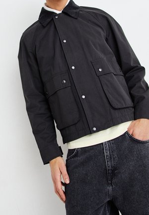 Mężczyzna w czarnej zapinanej na guziki kurtce z klapkami na kieszeniach i ciemnych dżinsach, z częściowo wsuniętą ręką do kieszeni, stojący na jednolitym tle.