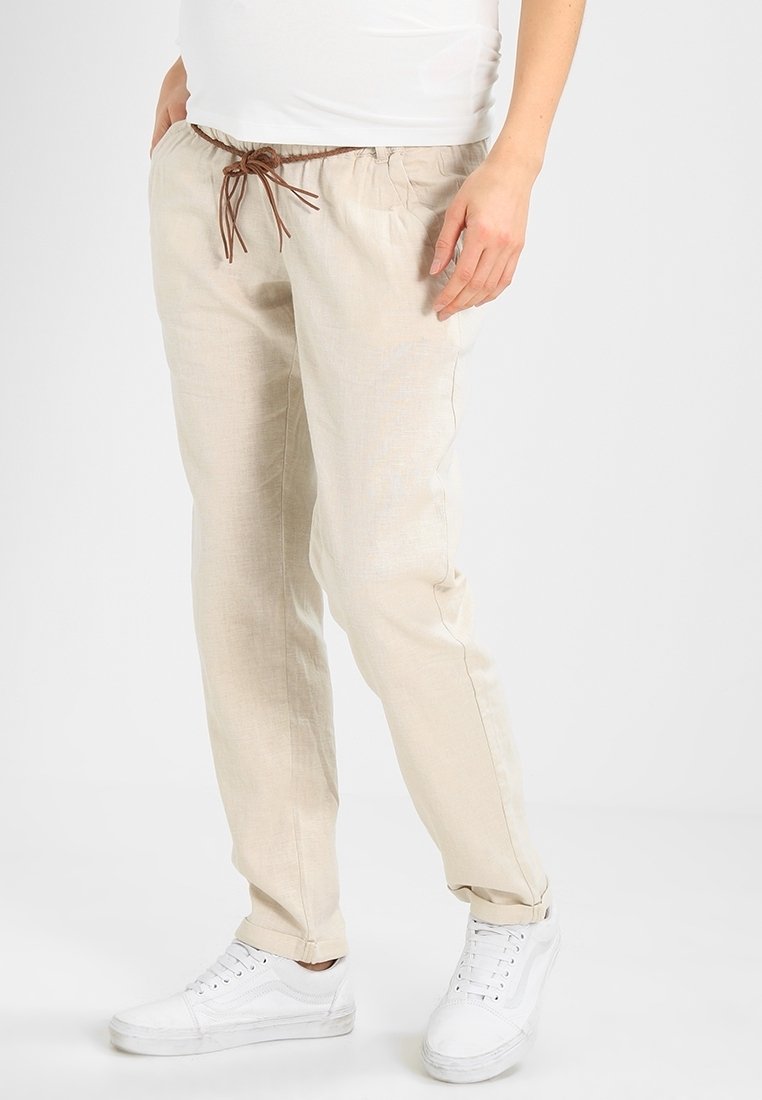 Světlé béžové lněné kalhoty s pružným pasem a uvolněným střihem. Hladká textura, v kombinaci s bílými teniskami, ukazuje neformální styl.