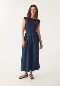 Marinblå klänning med kapärmar tillverkad av mjukt tyg, med en åtsittande bodice och en lös, samlad kjol. Matches med mörkbruna sandaler.