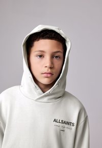 Sudadera con capucha gris claro con una capucha grande y suelta, que presenta el nombre de la marca "ALLSAINTS" impreso en negro en el lado izquierdo. Textura suave.