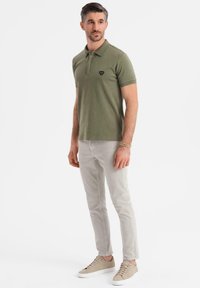 Olivgrön polo med kort ärm, prydd med en liten svart emblem. Ljust grå slim-fit byxor och beige sneakers med snören.