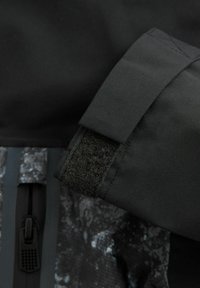 Giacca nera impermeabile con tessuto texturizzato, dotata di polsini in velcro, una tasca con zip e un pannello a motivo lungo il lato.