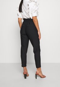 Svarta byxor med stickat mönster, åtsittande design, elastisk midja och avsmalnande ben. Tillsammans med en blus med volanger i vitt och sandaler med genomskinlig klack.