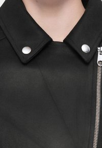 Giacca nera con colletto strutturato, bottoni a pressione argentati e zip asimmetrica. Texture morbida, design minimalista, senza motivi.