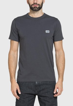 Uomo con barba curata che indossa una maglietta semplice a maniche corte grigio scuro con piccolo logo sul petto sinistro e jeans scuri su sfondo uniforme.
