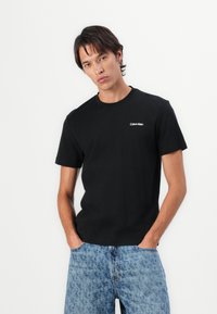 Schwarzes Baumwoll-T-Shirt mit Rundhalsausschnitt und kurzen Ärmeln; zeigt ein weißes "Calvin Klein"-Textlogo auf der Brust. Getragen mit blauen gemusterten Jeans.