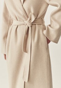Bež volneni plašč z zavezovalnim pasom, širokimi rokavi in dvema stranskima žepoma, z mehko teksturo in čistimi linijami.