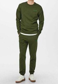 Bluza i spodnie dresowe w oliwkowej zieleni z miękkiego materiału, z okrągłym dekoltem i subtelnym logo w dopasowanym kolorze. Całość dopełniają białe trampki.