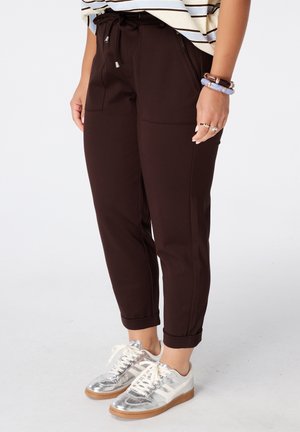 Persoon draagt donkerbruine omslagbroek met zakken, zilveren sneakers met witte veters en een gestreept topje, staand op een lichte vloer.