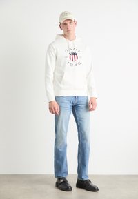Hvit hettegenser med logo og rød/blå skjold, kombinert med lyseblå jeans. Svart sko med spenner, står på et nøytralt gulv.