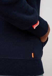 Felpa blu navy con polsini e orlo a coste, dotata di un interessante etichetta logo rossa sulla manica e una cucitura arancione accento sul lato.