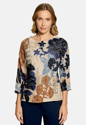 Femme portant un pull beige avec de grands motifs floraux bleus, noirs et bruns, associé à un pantalon bleu foncé, debout devant un fond uni.