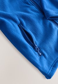 Detalje af en blå fleecejakke med en sidelomme med lynlås, glat tekstur og en tætsiddende pasform. Lynlåsen er blå med en tekstureret trækflig.