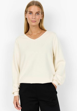 Cream V-neck pullover sweater with long sleeves, soft texture, and slightly relaxed fit, paired with black pants. Minimalistic design.