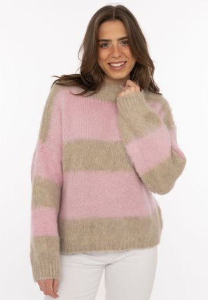 Junge Frau lächelnd, trägt einen weichen, flauschigen Pullover mit breiten horizontalen pinken und beigen Streifen, kombiniert mit weißen Hosen.