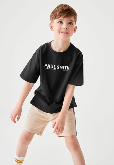 Melnas īsā piedurkne kokvilnas T-krekls ar baltu uzrakstu "PAUL SMITH". Kombinēts ar bežām šortiem un svītrainām zeķēm dzeltenā, baltā un pelēkā krāsā.