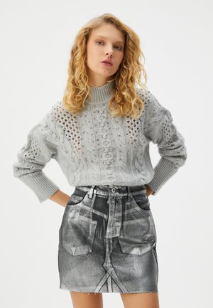 Maglione grigio chiaro lavorato a maglia con cavi e dettagli traforati, abbinato a una gonna di jeans nera a vita alta con un motivo sbiadito e orlo sfilacciato.