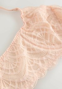 Soutien-gorge bralette en dentelle rose clair avec motifs floraux et géométriques, fines bretelles ajustables et bords festonnés sur un fond uni.