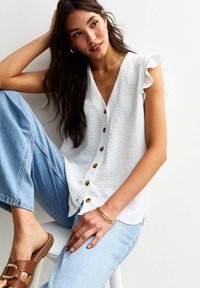 Vit texturerad blus med V-ringning och volangärmar, med guldknappar framtill. Bärs med blå denimjeans och bruna sandaler.