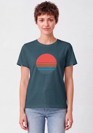 Persoon met kort bruin haar met een donker turquoise t-shirt met een rood-blauw gestreept cirkeldesign en lichtblauwe spijkerbroek.