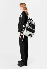 Mochila acolchada en negro y gris con múltiples bolsillos y cremalleras, con parche de marca y correas ajustables. Superficie texturizada.