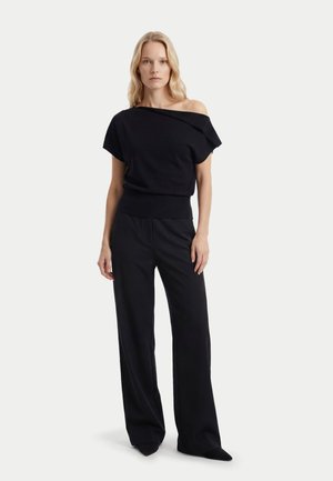 Frau mit blondem Haar steht und trägt ein schwarzes Off-Shoulder-Top und weite schwarze Hosen vor einem einfarbigen weißen Hintergrund.