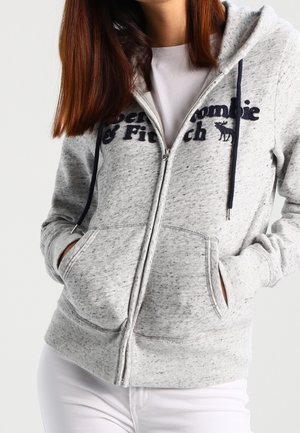 Grå zip-up hoodie lavet af blødt stof, med et frontlogo, to lommer og snore i hætten.