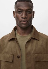 Veste en suédine marron avec une fermeture à bouton, une poche poitrine et une texture lisse, portée sur un t-shirt vert à col rond.