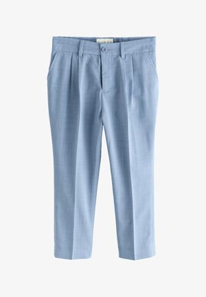 Junge trägt einen hellblauen Anzug, weißes Hemd, blaue Fliege und weiße Sneaker, steht vor einem schlichten hellen Hintergrund und lächelt.