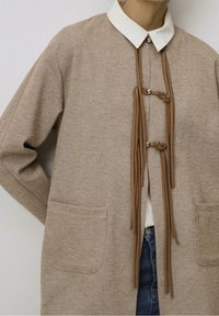 Beżowy, teksturowany płaszcz z białym kołnierzem, ozdobiony brązowymi zawiązanymi trokami i dwoma przednimi kieszeniami. Prosty, minimalistyczny krój z luźnym dopasowaniem.