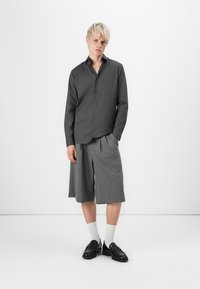 Camicia grigia con collo a punta, dalla vestibilità slim, abbinata a pantaloni corti grigi con pieghe. Il modello indossa calze bianche e mocassini neri. Tessuto dalla superficie liscia.